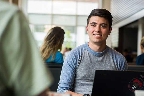 A man smiles while sitting at a table with a laptop